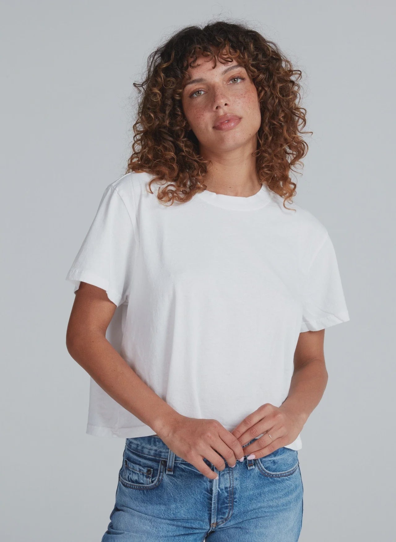 A person with curly brown hair wears the ASKK Shrunken Boy Tee and blue jeans, standing against a light gray background. They face the camera with a slight smile, hands loosely clasped.