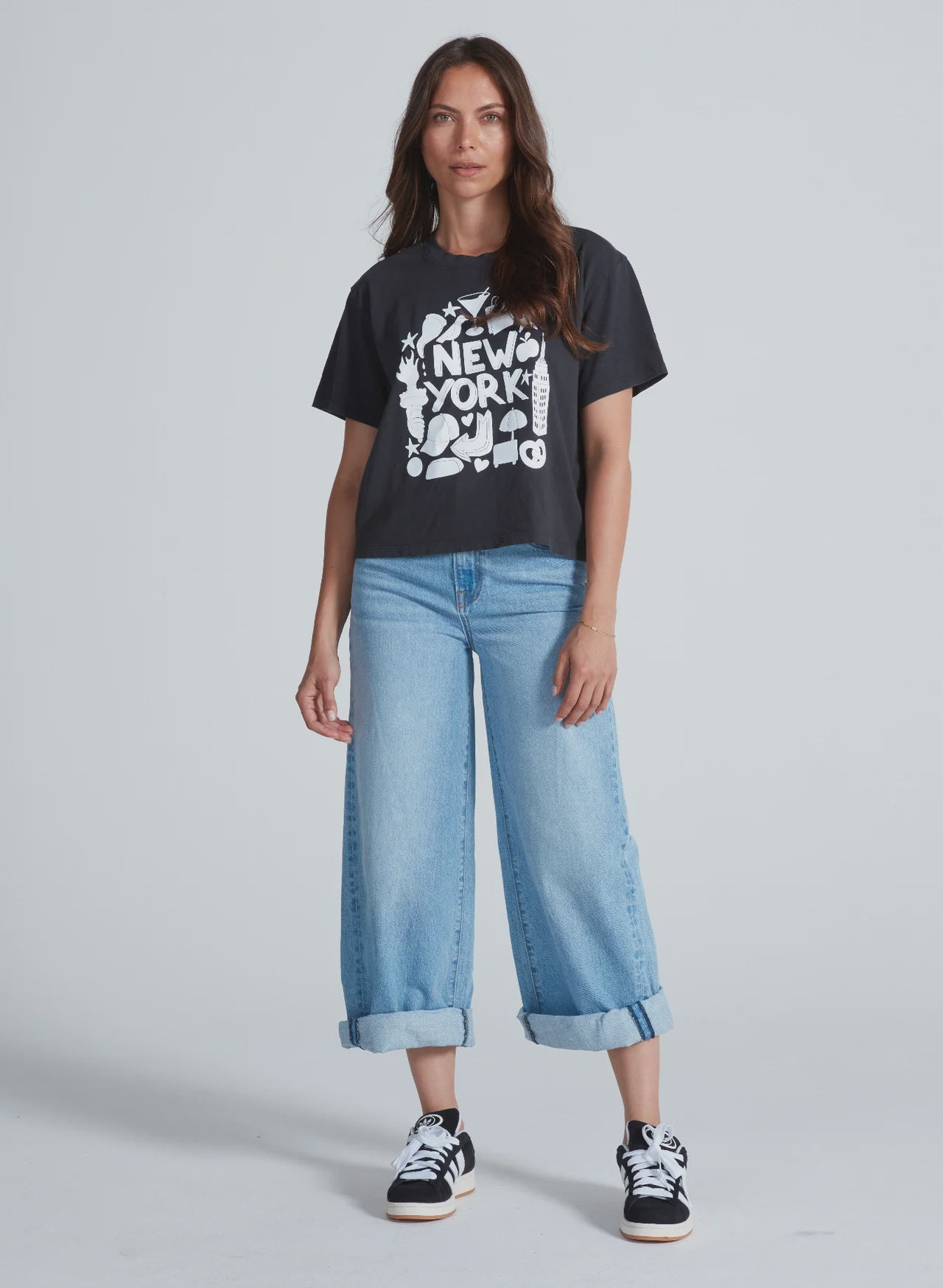 A woman faces the camera in an ASKK Shrunken Boy Tee—a black "New York" crewneck—paired with light blue wide-leg jeans with rolled cuffs and black sneakers with white soles, set against a plain light background.