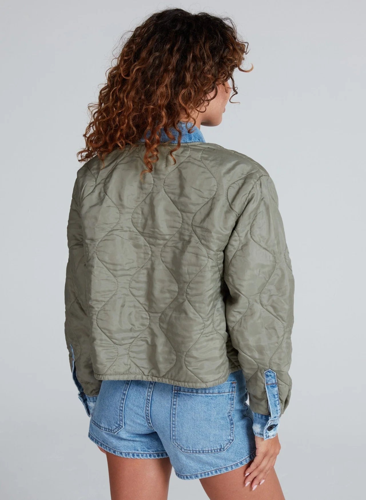 A person with curly brown hair is shown from behind, wearing The Peak Liner green cropped quilted jacket by ASKK over a blue denim shirt and denim shorts against a light gray background.