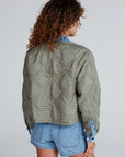 A person with curly brown hair is shown from behind, wearing The Peak Liner green cropped quilted jacket by ASKK over a blue denim shirt and denim shorts against a light gray background.