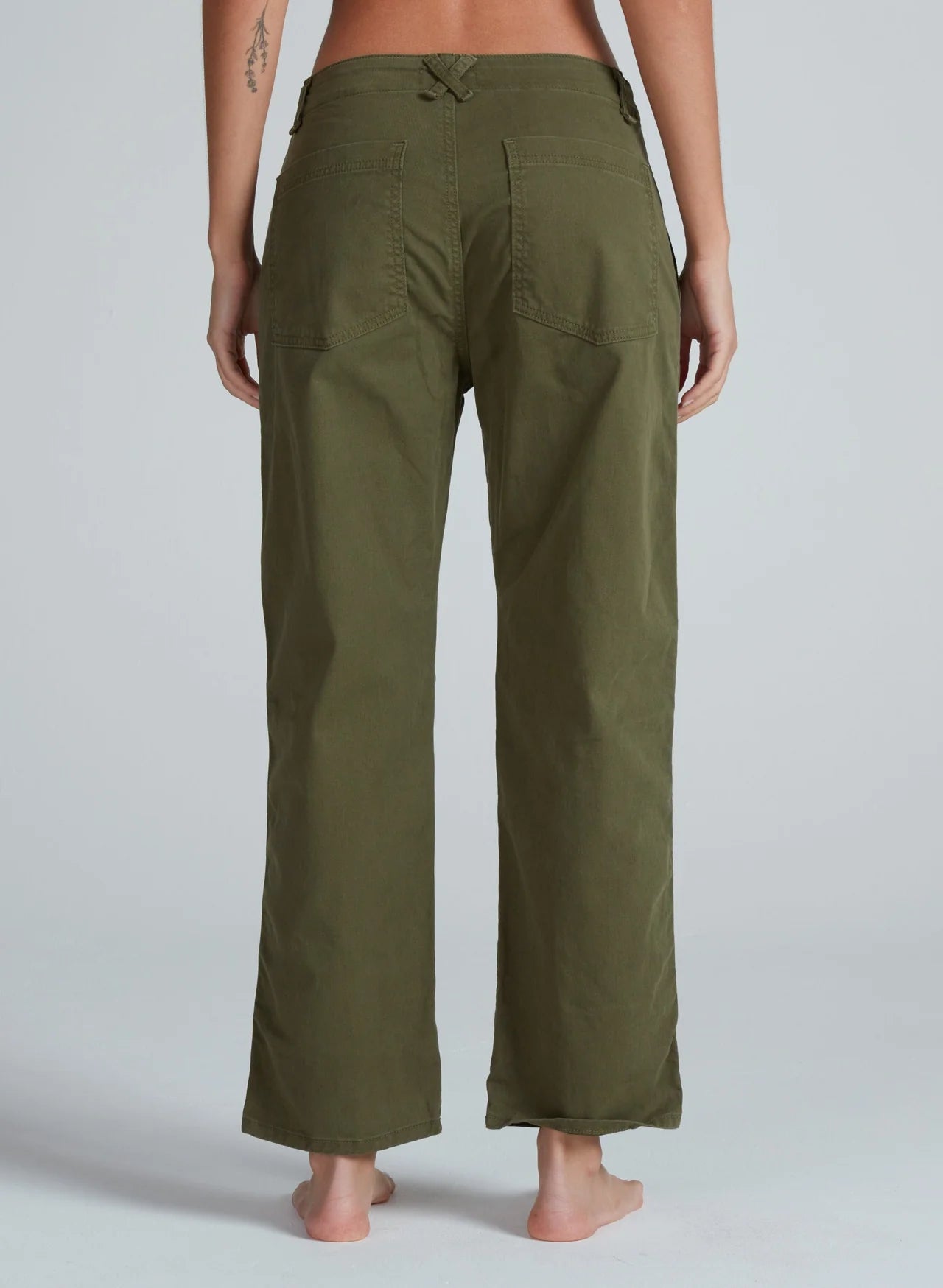 A person stands barefoot, facing away from the camera, wearing ASKK’s Tomboy Chino in olive green—a mid-weight pant with large back pockets and a detailed waistband. The background is plain and light-colored.