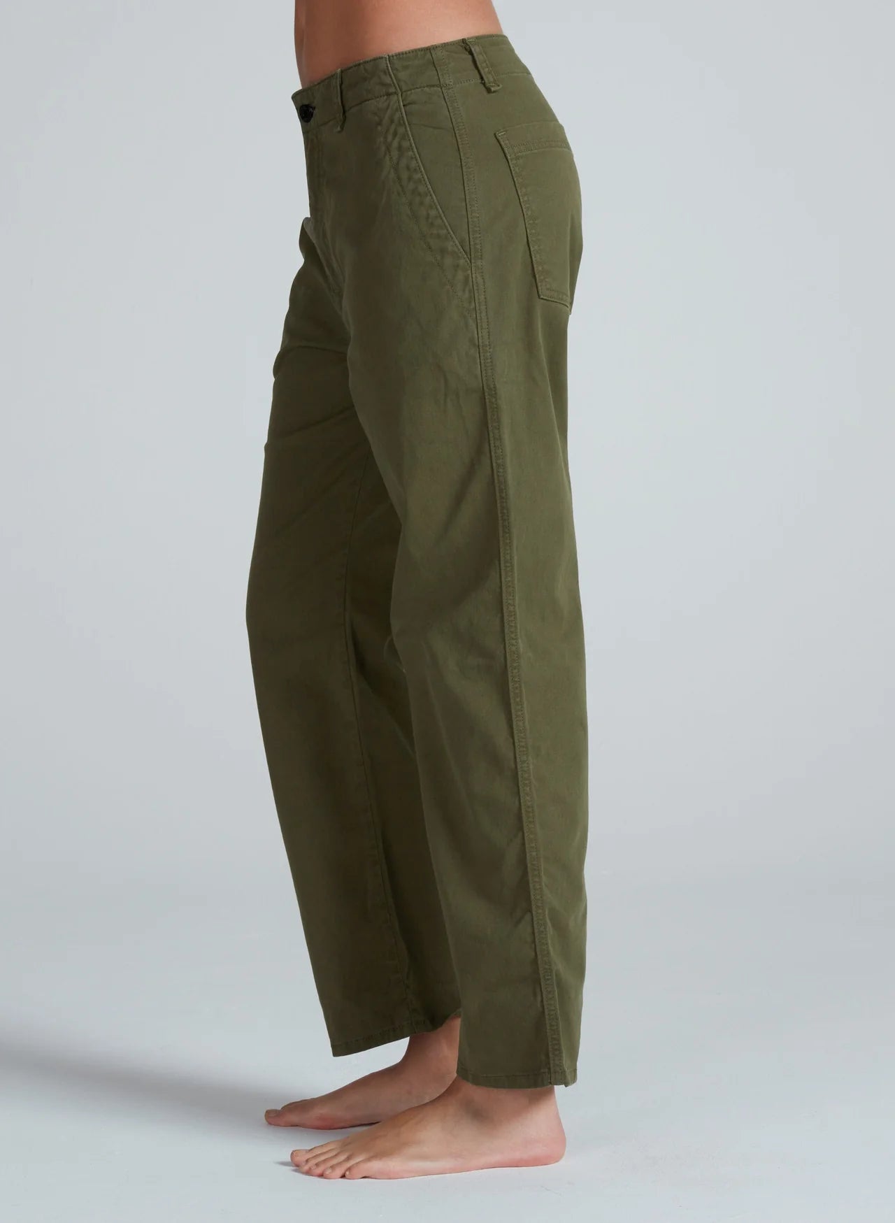 A person stands barefoot against a plain light gray background, wearing ASKK's high-waisted Tomboy Chino in olive green with visible side and back pockets. The loose-fitting, midweight pants are photographed from the side.