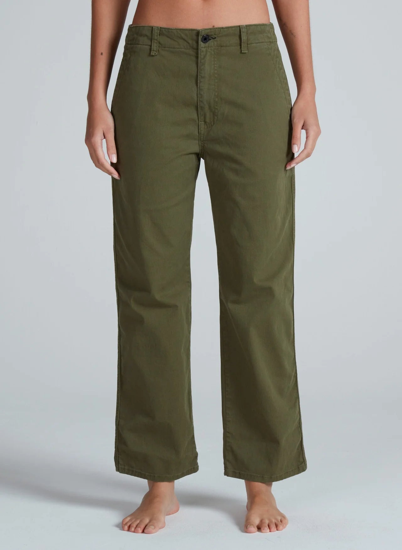A person stands barefoot, wearing ASKK's Tomboy Chino—high waist, straight-leg olive green pants made from stretchy twill. The waist-down shot highlights these mid weight chinos against a plain light gray background.