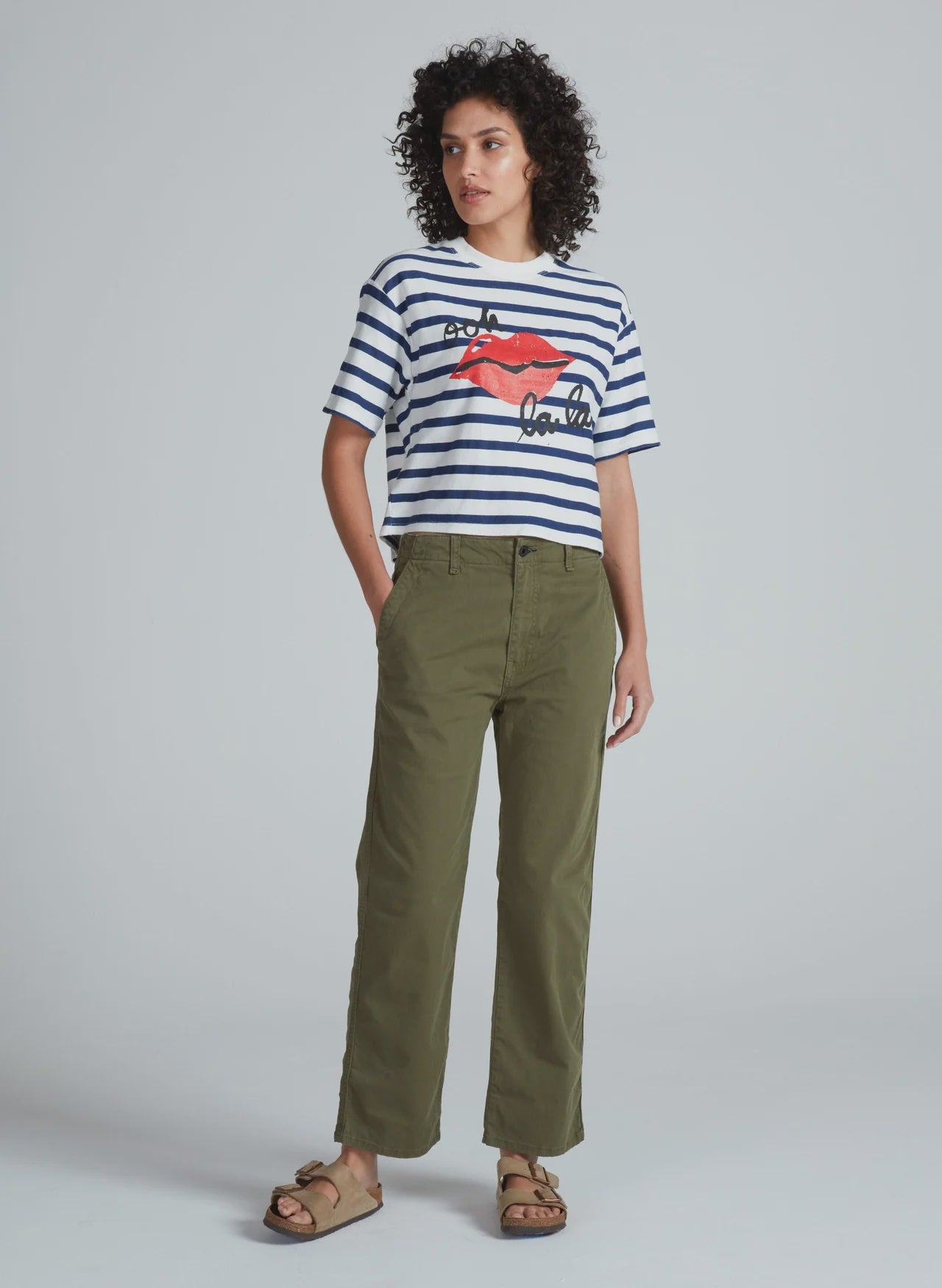 A person stands against a plain background wearing a striped short-sleeve shirt with a red graphic, tan sandals, and ASKK's Tomboy Chino high waist olive green stretchy twill pants with hands in pockets.