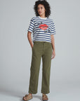 A person stands against a plain background wearing a striped short-sleeve shirt with a red graphic, tan sandals, and ASKK's Tomboy Chino high waist olive green stretchy twill pants with hands in pockets.