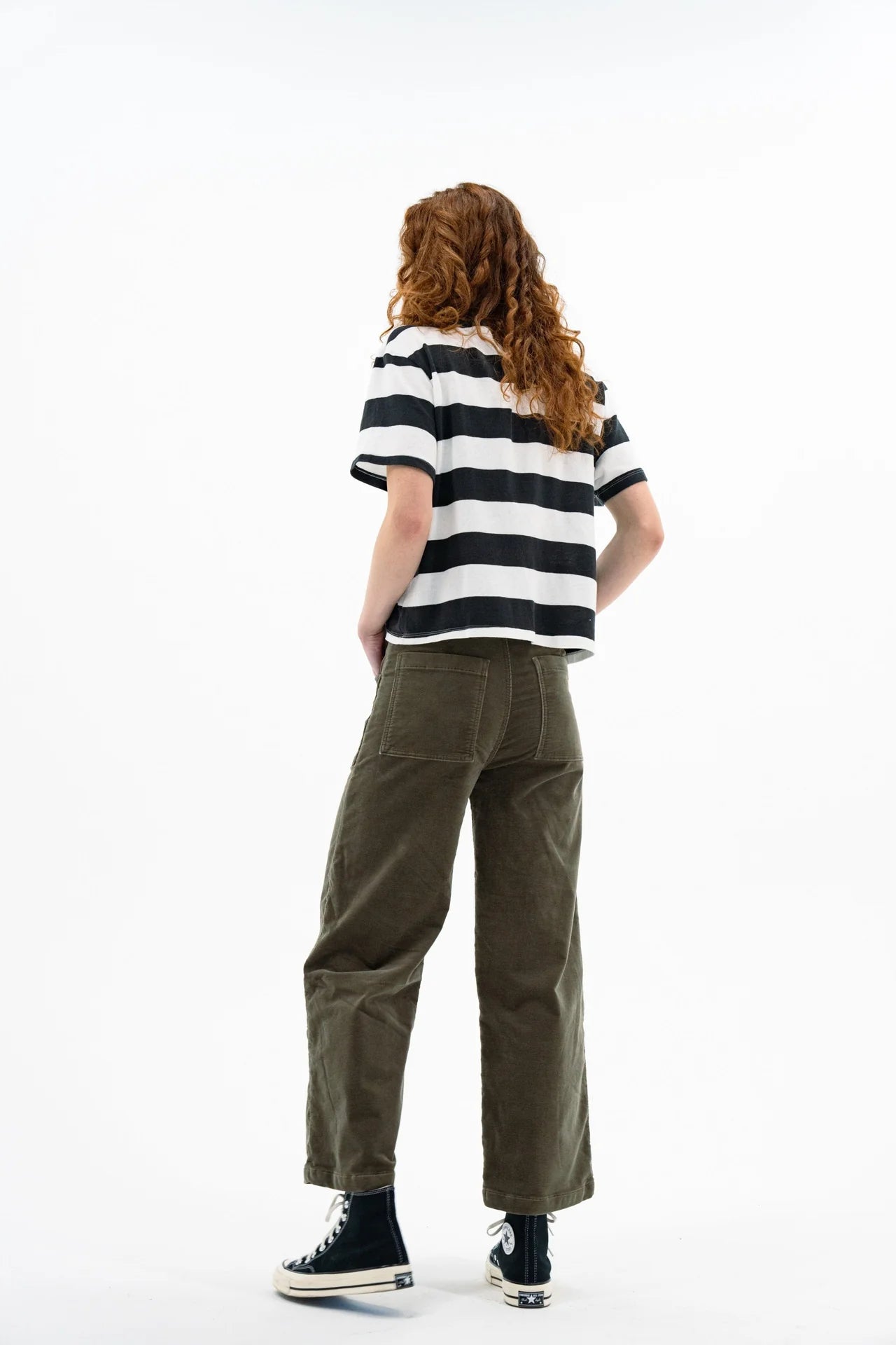 A person with long curly hair, wearing a black and white striped t-shirt and ASKK's Virginia Corduroy high-rise wide-leg pants, stands facing away from the camera against a plain white background.
