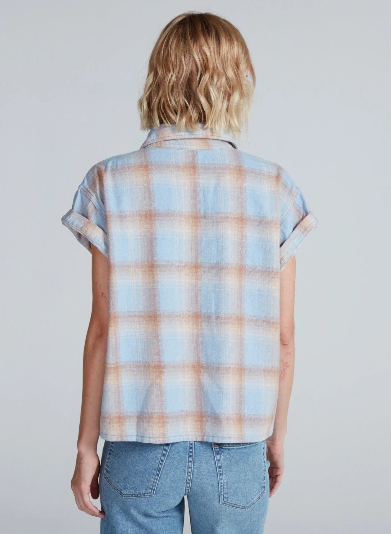 A person with short, wavy blonde hair is shown from the back, wearing the ASKK Virginia Shirt—a relaxed short-sleeve button-up plaid made of lightweight cotton—paired with light blue jeans.