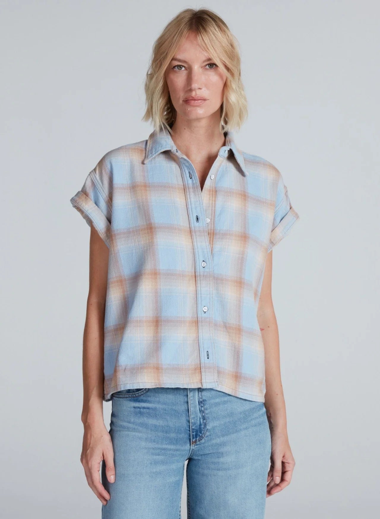 A woman with blonde, shoulder-length hair models the ASKK Virginia Shirt—a relaxed, short-sleeve button-up in pastel plaid lightweight cotton—paired with light blue jeans, standing against a plain light gray backdrop.