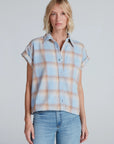 A woman with blonde, shoulder-length hair models the ASKK Virginia Shirt—a relaxed, short-sleeve button-up in pastel plaid lightweight cotton—paired with light blue jeans, standing against a plain light gray backdrop.