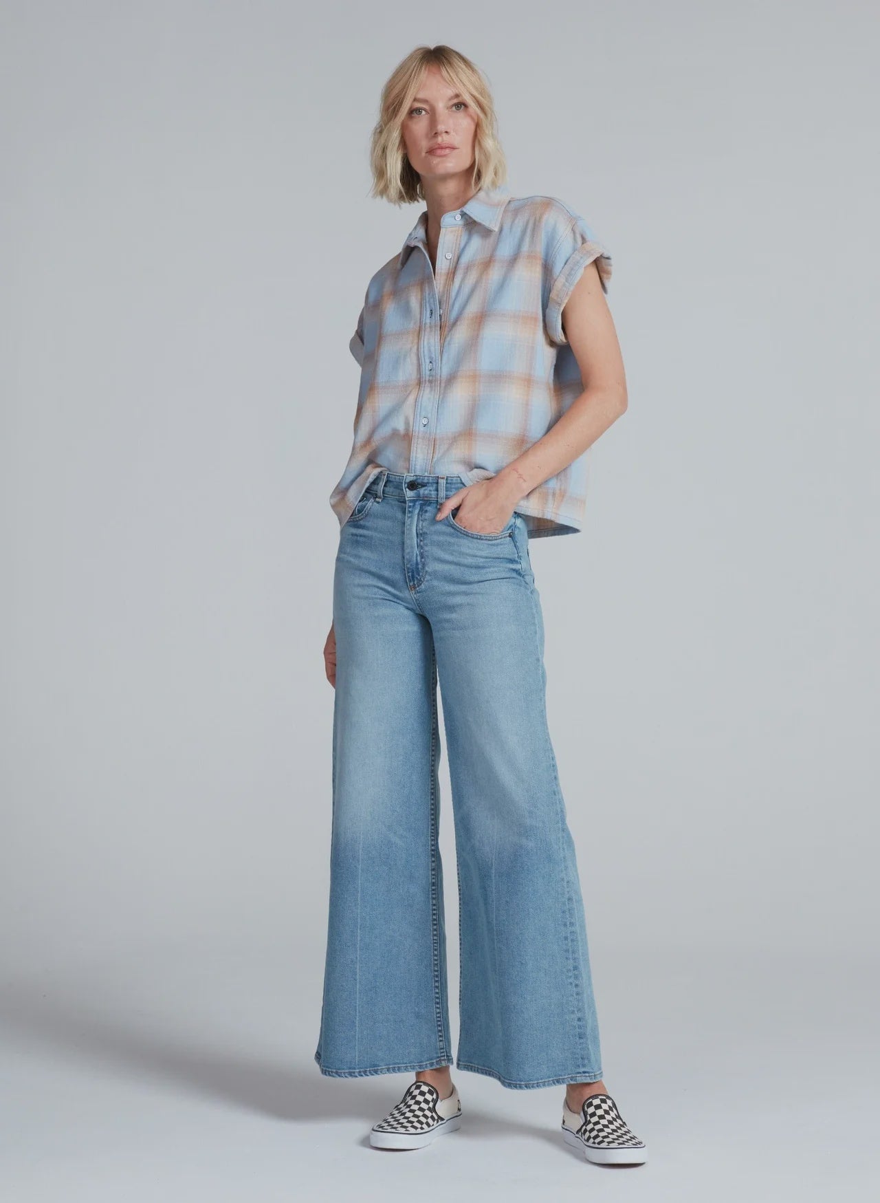 Wearing the ASKK Virginia Shirt, this person stands against a plain background in a relaxed pose with one hand in their pocket, paired with high-waisted wide-leg jeans and checkered slip-on sneakers.