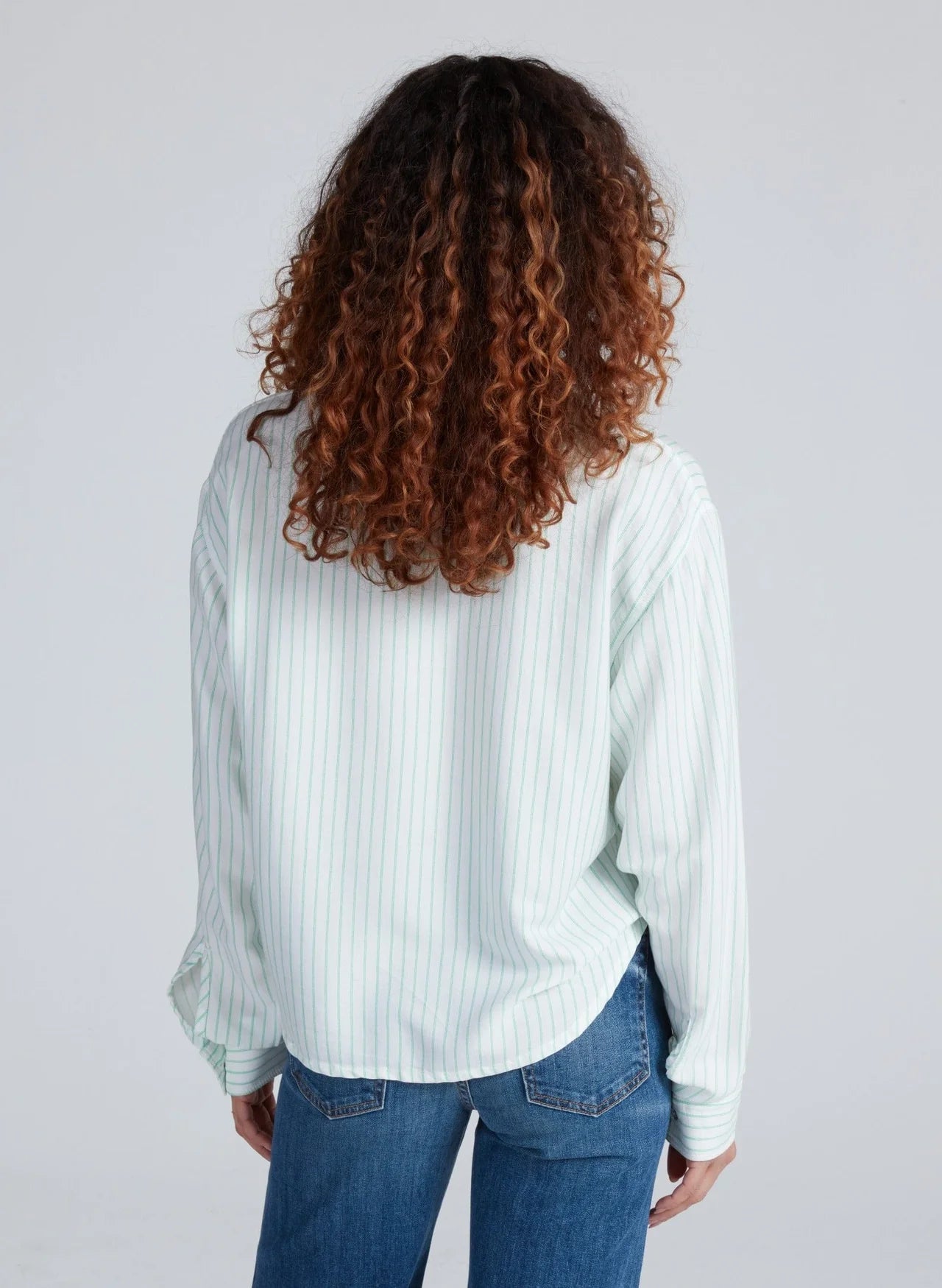 A person with curly brown hair is shown from the back wearing the ASKK Crosby Shirt—a relaxed-fit, light pinstriped long-sleeve shirt—with blue jeans, set against a plain light background.