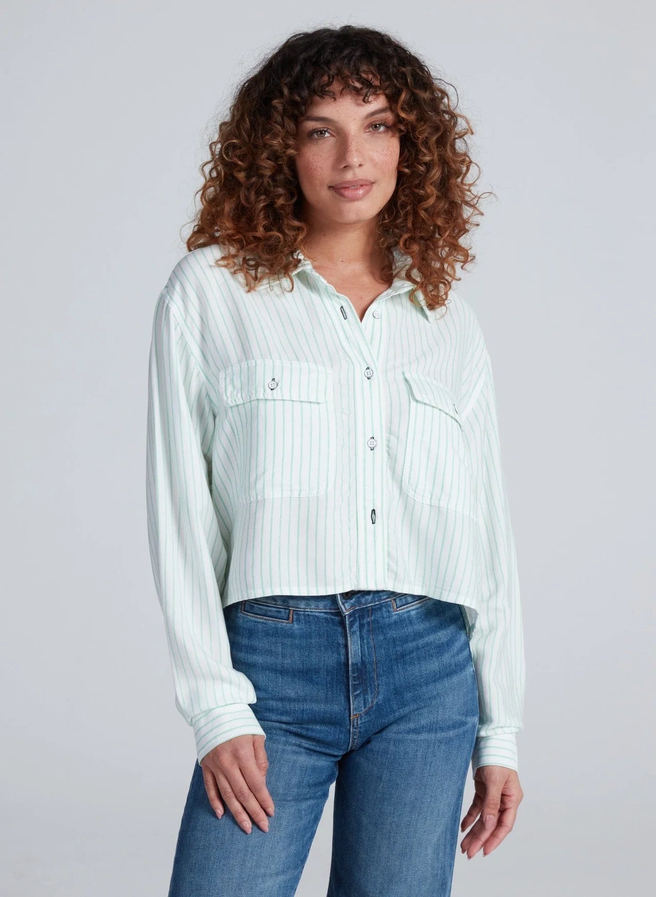 A woman with curly brown hair wears the ASKK Crosby Shirt, featuring light green and white stripes and chest pockets, paired with blue jeans, standing against a plain light background.