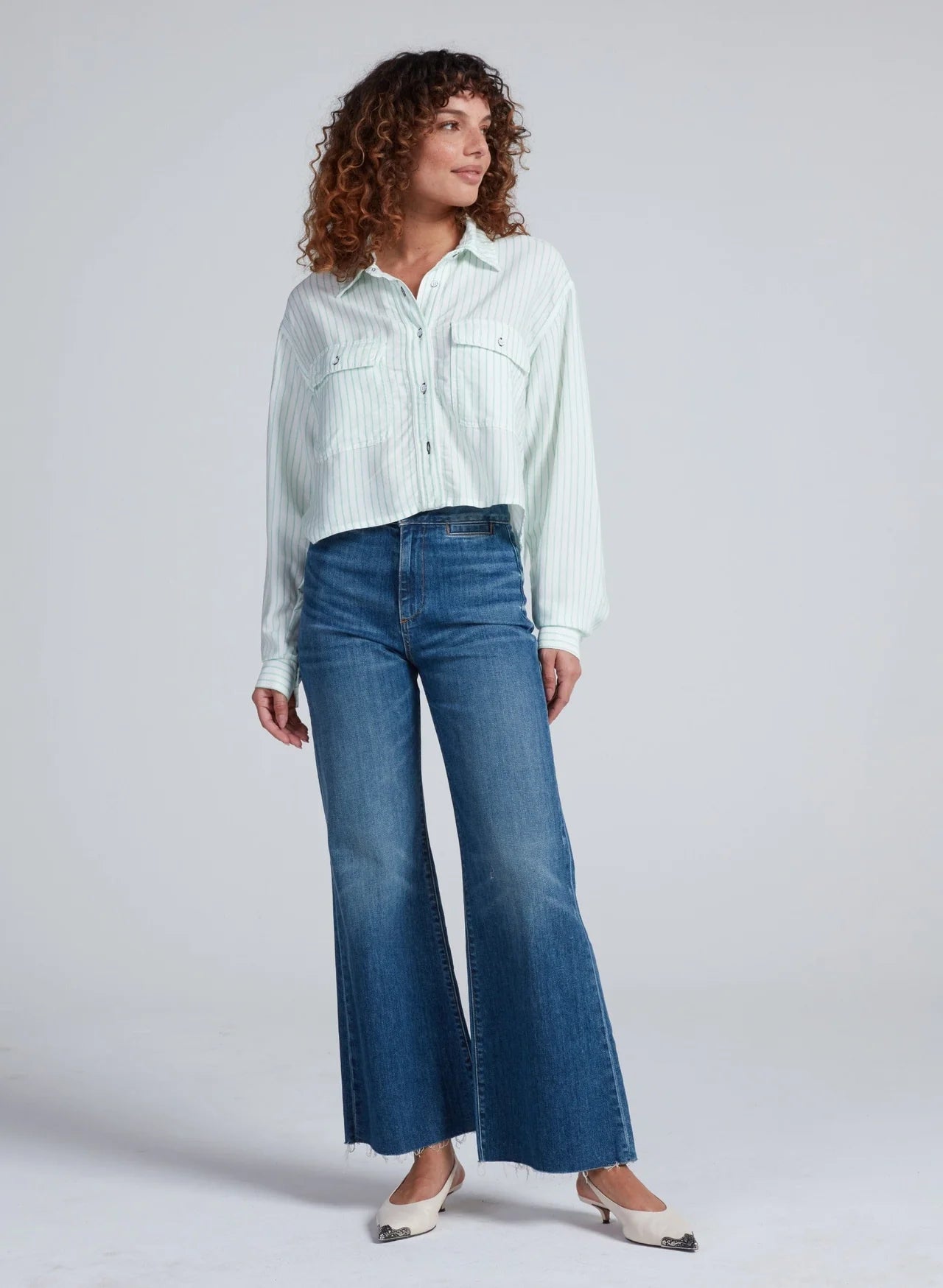 Wearing the ASKK Crosby Shirt, a woman with curly hair pairs it with blue high-waisted wide-leg jeans and cream flats, standing against a plain light background.