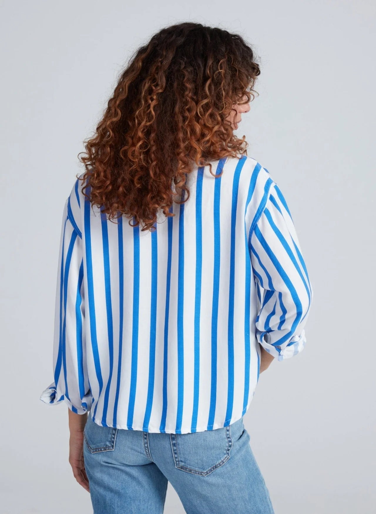 A person with curly brown hair, seen from behind, wears the ASKK Crosby Shirt—a blue and white vertically striped 100% cotton relaxed fit—and light blue jeans against a plain light background.