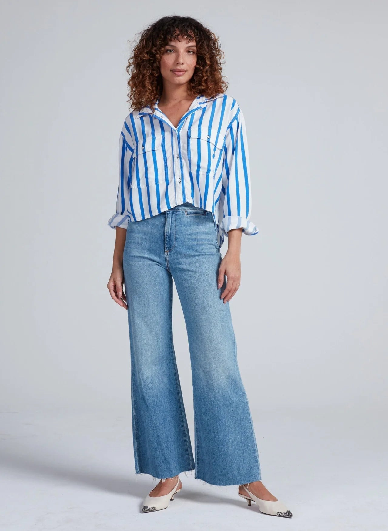 A person wears the ASKK Crosby Shirt, a relaxed fit blue and white vertically striped 100% cotton button-up, paired with light blue flared jeans and white shoes, against a plain background.