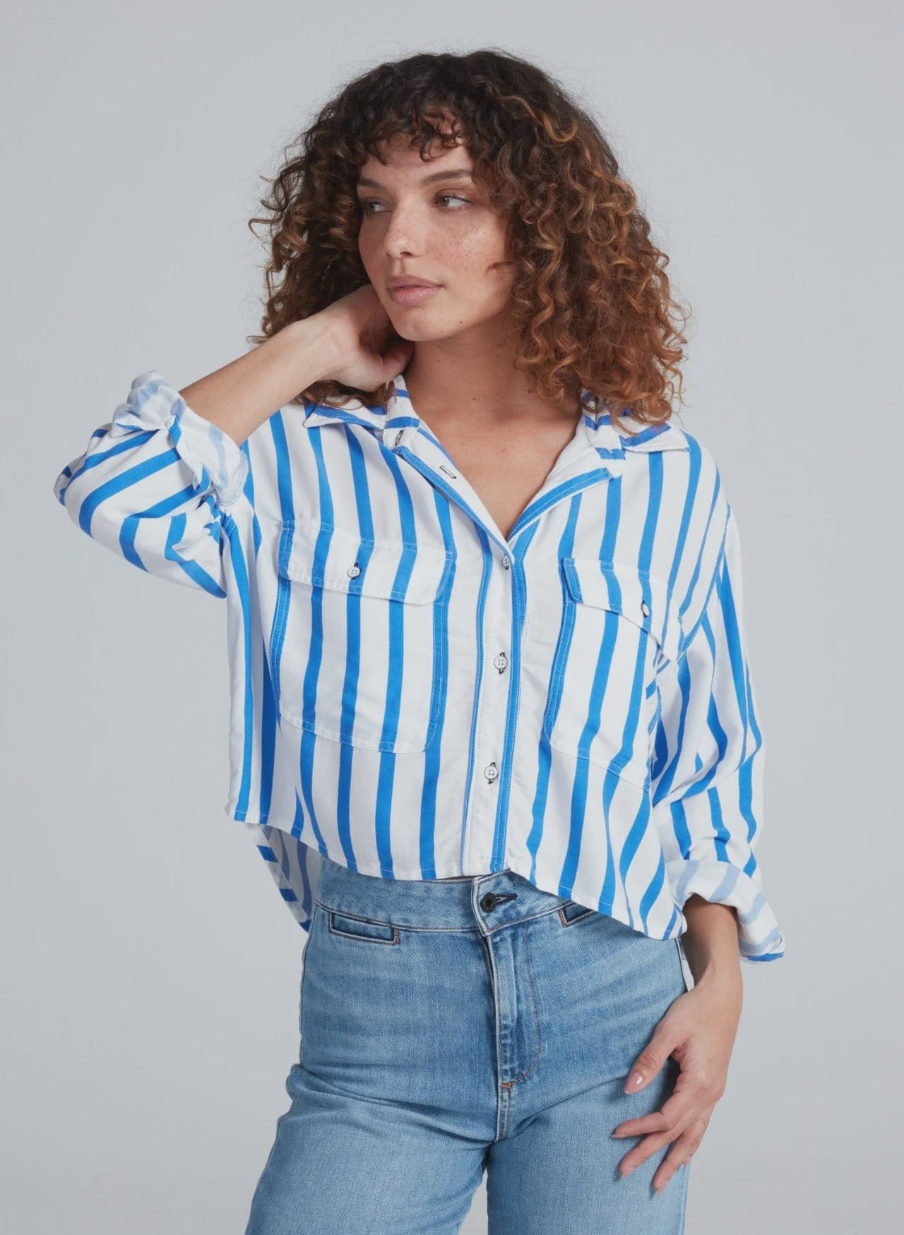 A person with curly brown hair wears the ASKK Crosby Shirt—blue and white vertical stripes, 100% cotton, relaxed fit—and light blue jeans, posing with one hand touching their neck against a plain light background.