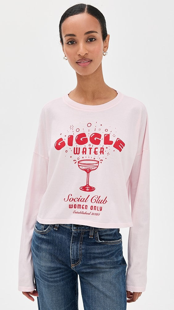 A woman wears the ASKK Cropped LS Tee, a light pink long-sleeve shirt made from 100% cotton with red “GIGGLE WATER Social Club WOMEN ONLY Established 2020” text and a cocktail glass design, standing before a plain white background.