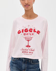 A woman wears the ASKK Cropped LS Tee, a light pink long-sleeve shirt made from 100% cotton with red “GIGGLE WATER Social Club WOMEN ONLY Established 2020” text and a cocktail glass design, standing before a plain white background.