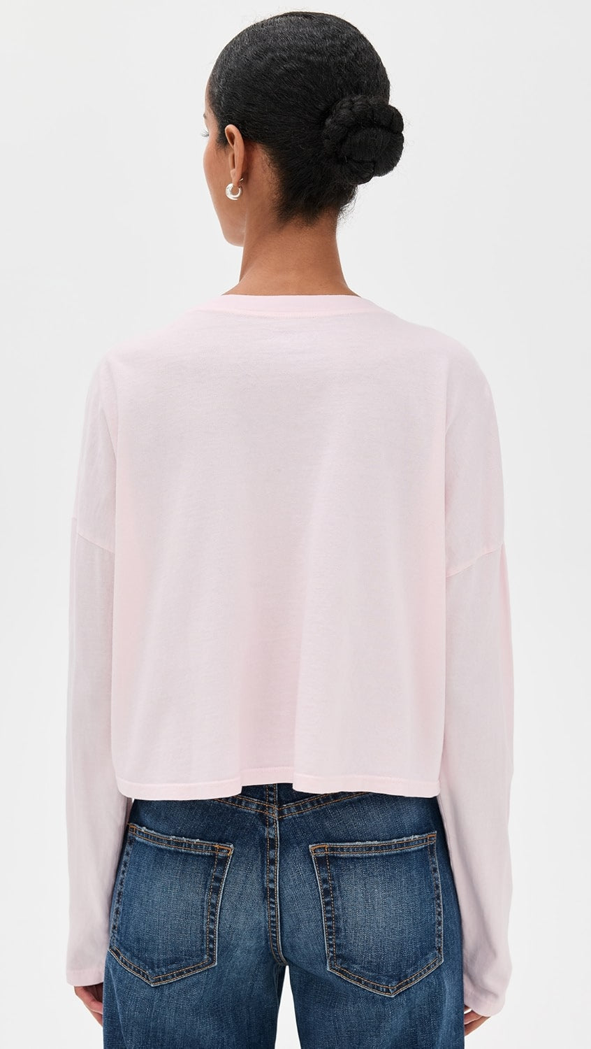 A person with dark hair in a neat bun, wearing small hoop earrings, the ASKK Cropped LS Tee in light pink, and blue jeans, is shown from the back against a plain white background.