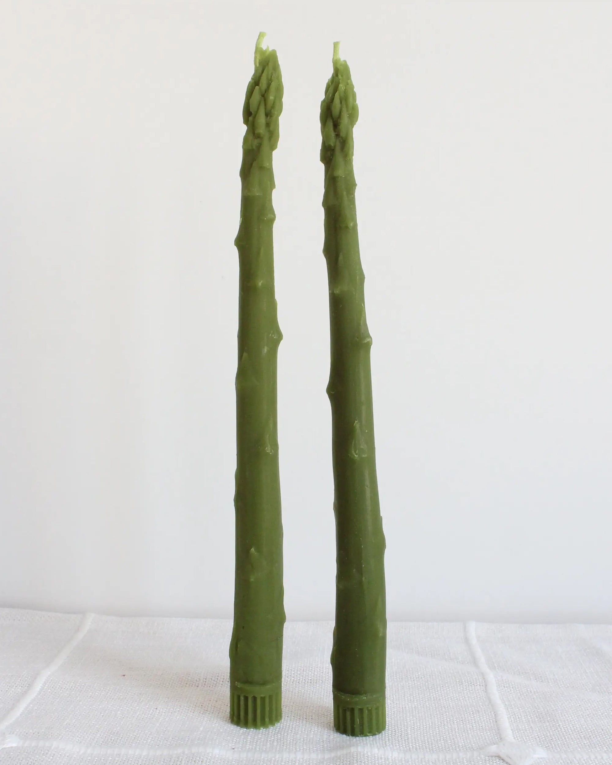 Two Faire Asparagus Beeswax Candles, shaped like green asparagus stalks, stand upright on a white textured surface against a plain white background.