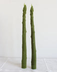 Two Faire Asparagus Beeswax Candles, shaped like green asparagus stalks, stand upright on a white textured surface against a plain white background.