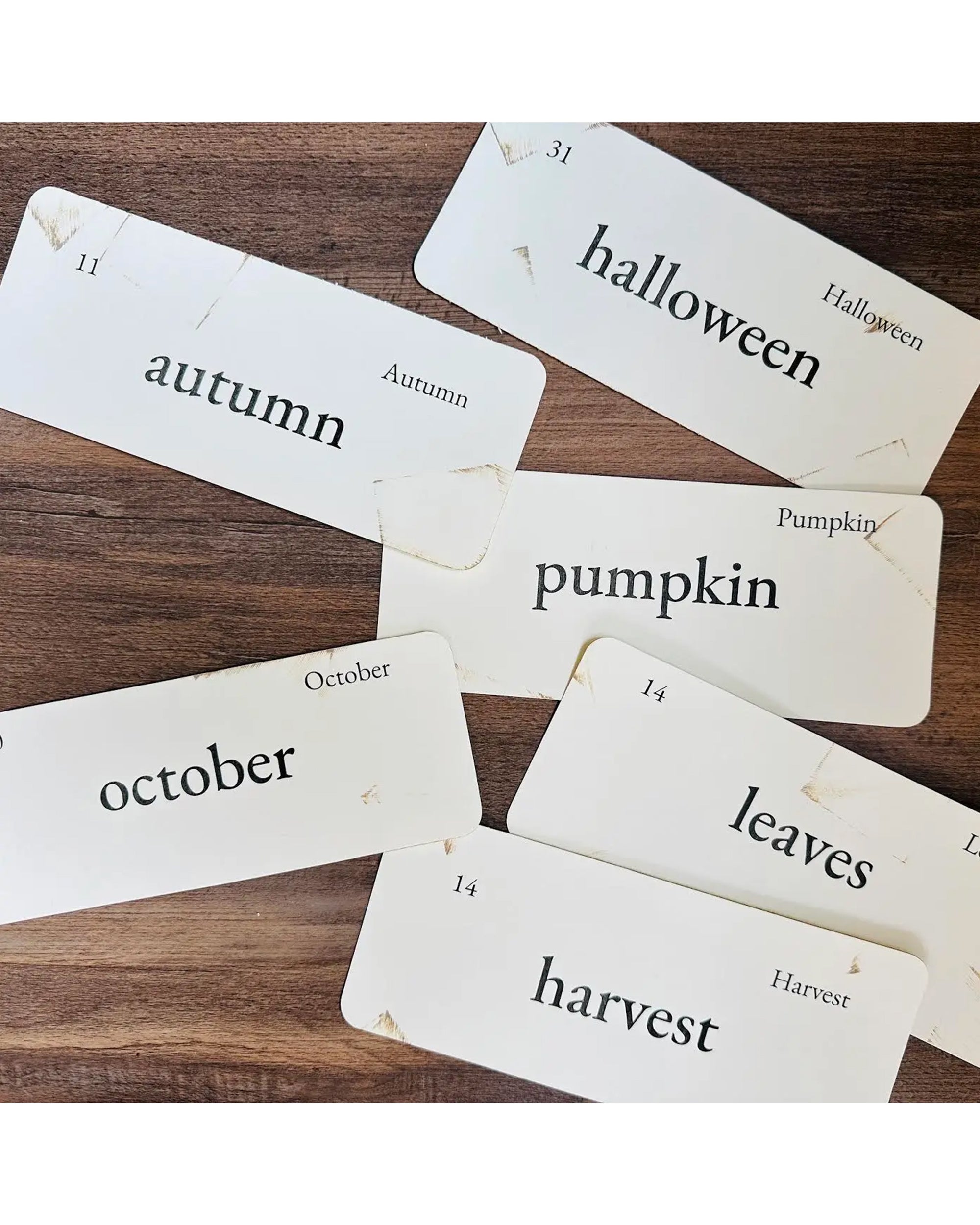 Six white Autumn Flashcards by Faire, each with black text featuring words like autumn, halloween, pumpkin, october, leaves, and harvest, are spread on a wooden surface—perfect seasonal decor.