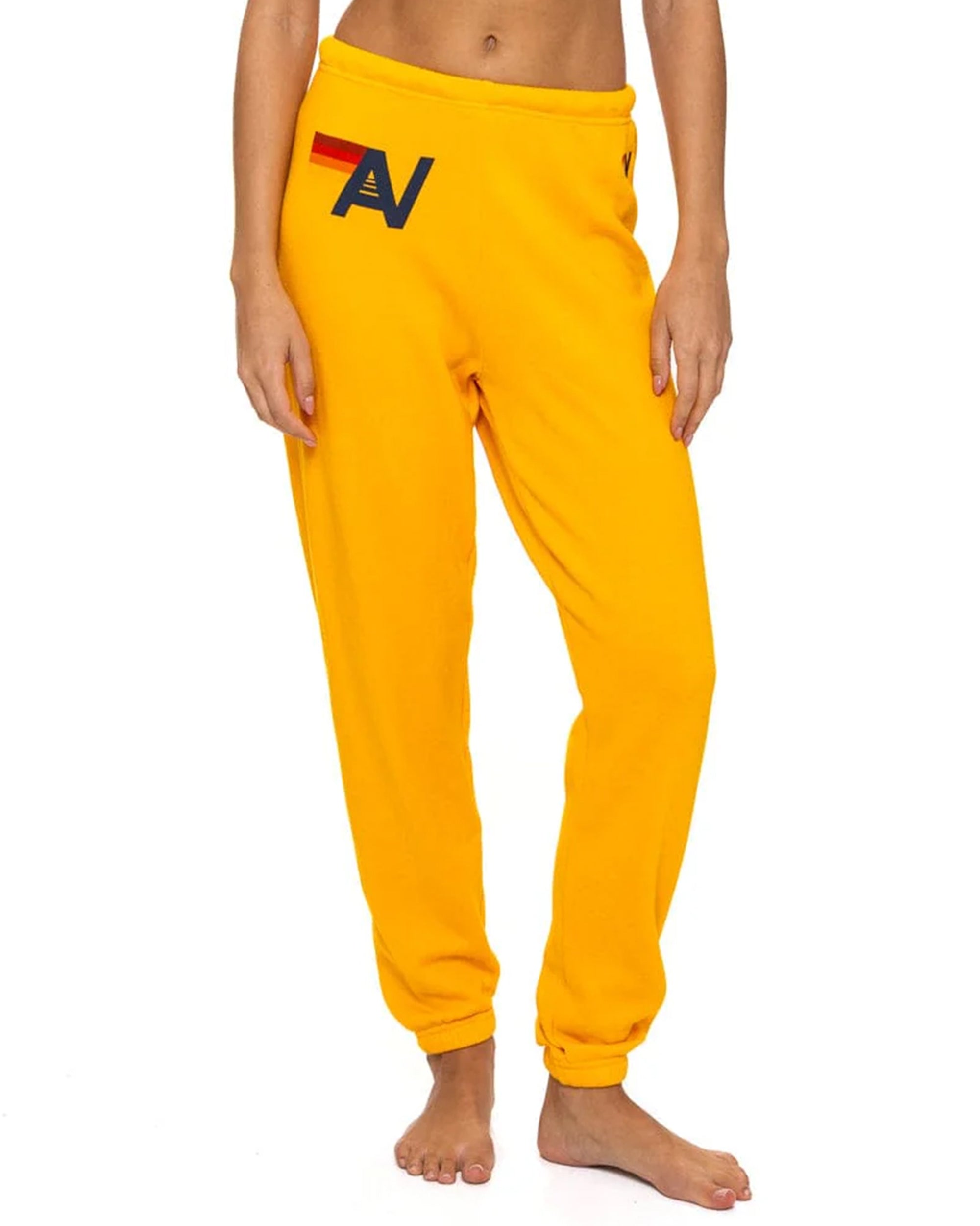 A model showcases Aviator Nation’s logo - womens sweatpants in bright yellow, featuring a red and black geometric "A" on the left hip. Only her lower torso and legs are visible as she stands barefoot against a white backdrop.
