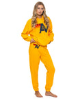 A woman stands barefoot against a white background, wearing an Aviator Nation logo - pullover hoodie relaxed in bright yellow with matching sweatpants, both featuring a bold "AV" logo and red stripes.