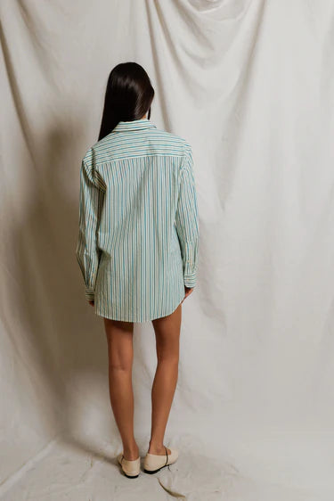 A person with long dark hair, wearing the Perfectwhitetee Avalon Poplin Button Down in blue and white stripes and white slip-on shoes, stands with their back to the camera against a light draped backdrop.