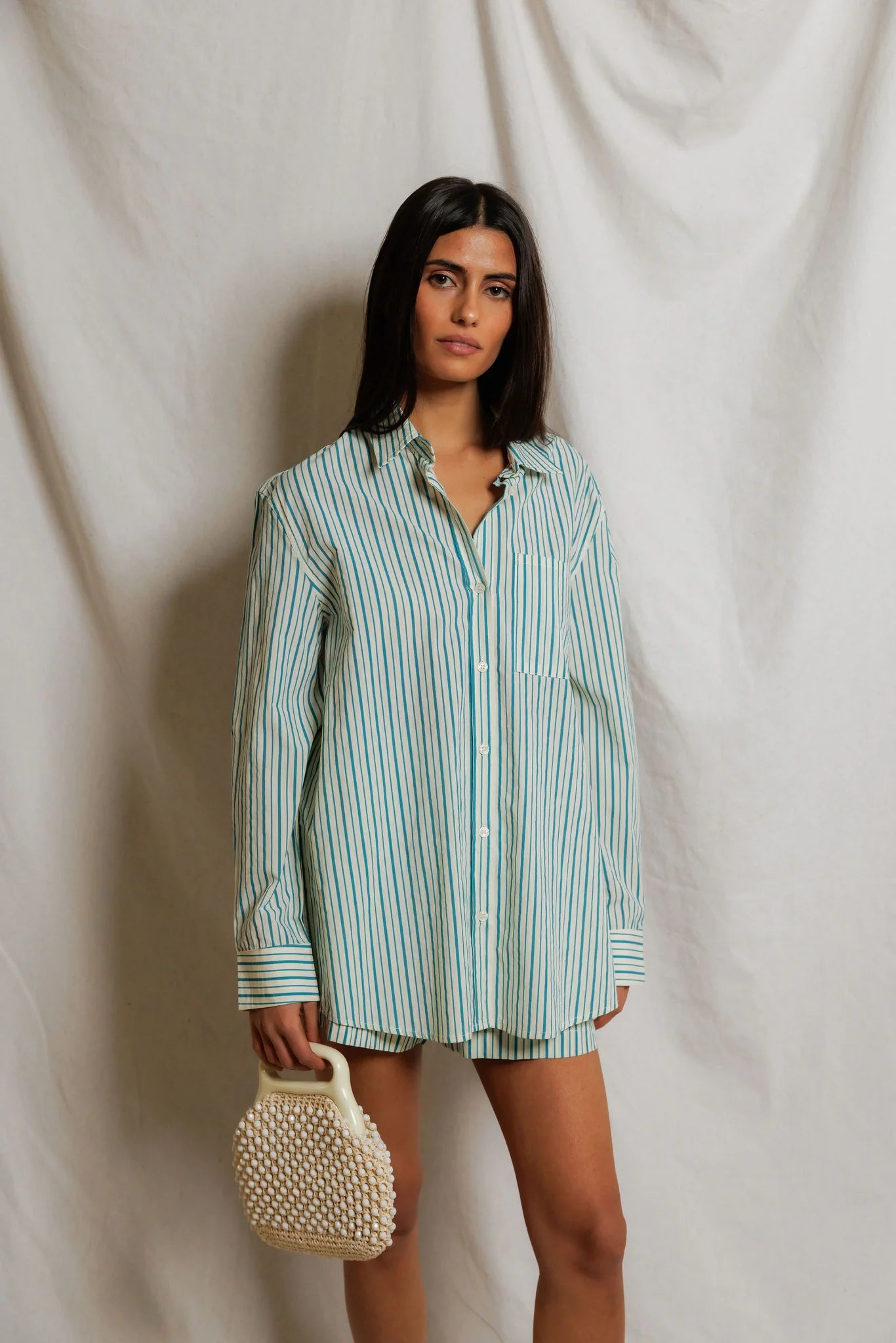 A woman stands before a white draped backdrop, wearing the Avalon Poplin Button Down by Perfectwhitetee with matching shorts. She holds a beaded handbag in her left hand and gazes toward the camera.