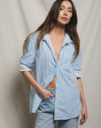 A woman with long brown hair wears the Perfectwhitetee Avalon Poplin Button Down, an oversized blue-and-white striped cotton shirt over a bare chest and light blue jeans, standing against a draped neutral fabric background.