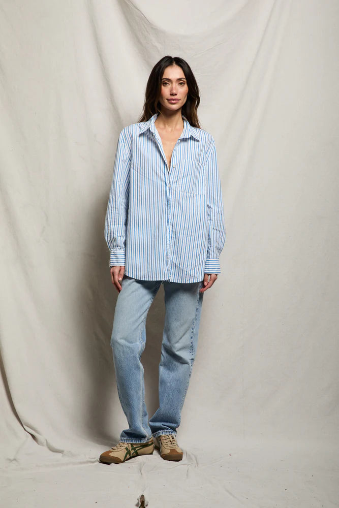 A woman stands before a beige draped backdrop, wearing the Avalon Poplin Button Down by Perfectwhitetee in blue and white stripes, paired with light blue jeans and tan sneakers. She looks at the camera with a relaxed expression.