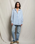 A woman stands before a beige draped backdrop, wearing the Avalon Poplin Button Down by Perfectwhitetee in blue and white stripes, paired with light blue jeans and tan sneakers. She looks at the camera with a relaxed expression.