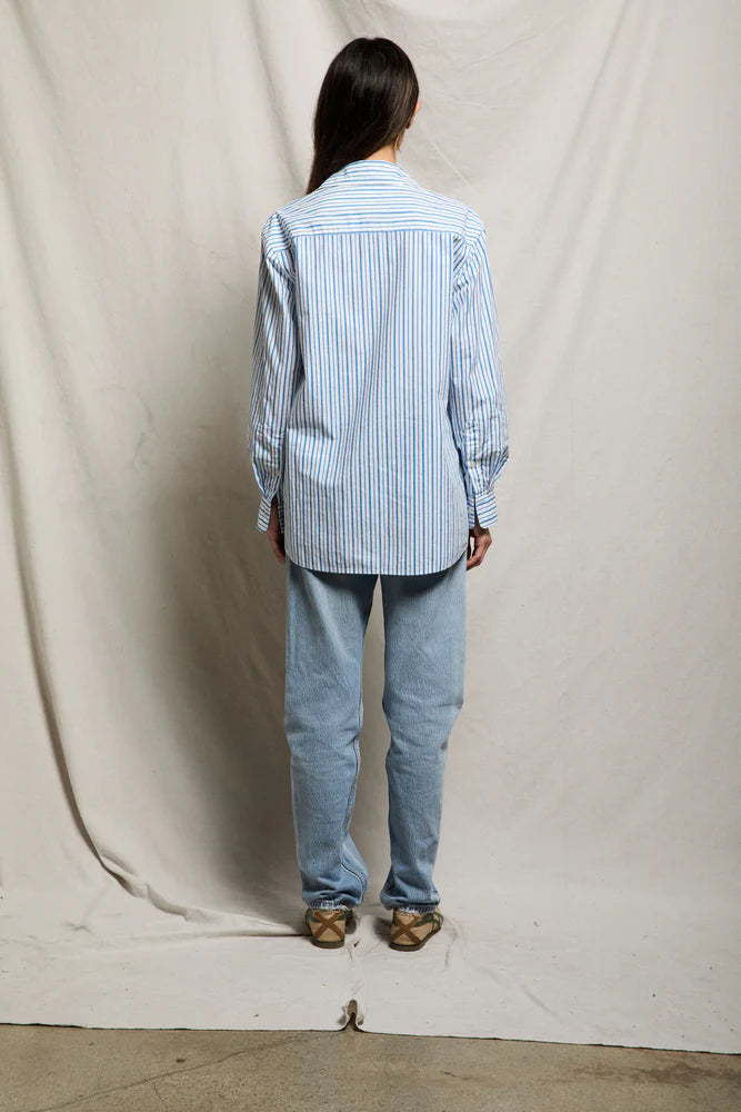 A person with long dark hair faces away, wearing the Avalon Poplin Button Down by Perfectwhitetee in blue and white stripes, paired with light blue jeans and beige shoes. Set against draped light fabric—an ideal chic summer cover-up look.
