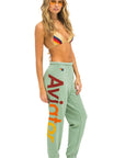 A woman with wavy blonde hair wears orange sunglasses, a colorful bikini top, and comfortable green sweatpants called "Aviator Nation 2 - womens sweatpants" by Aviator Nation with the word “Aviator” written down the side in red and yellow letters, giving a vintage feel. She stands barefoot against a white background.