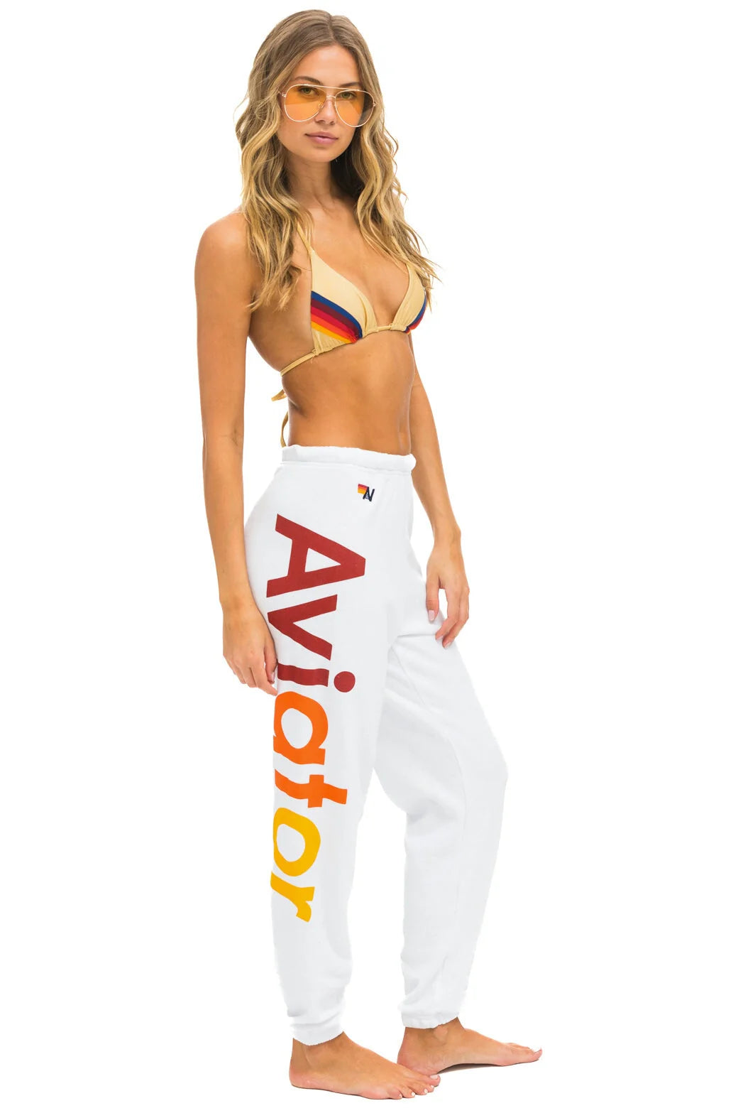 A woman stands barefoot, wearing comfortable white Aviator Nation 2 - Womens Sweatpants with the word &quot;Aviator&quot; in large red and yellow gradient letters on the side. She also has on a multicolored bikini top and yellow tinted sunglasses, with loose, wavy hair cascading over her shoulders, exuding a vintage feel.