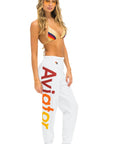 A woman stands barefoot, wearing comfortable white Aviator Nation 2 - Womens Sweatpants with the word "Aviator" in large red and yellow gradient letters on the side. She also has on a multicolored bikini top and yellow tinted sunglasses, with loose, wavy hair cascading over her shoulders, exuding a vintage feel.