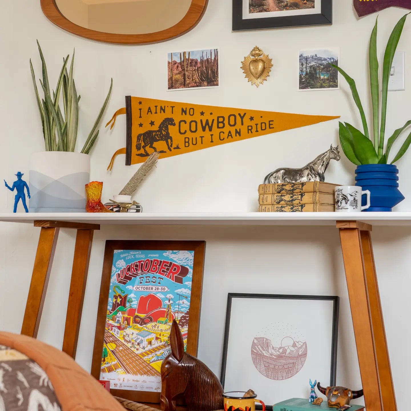 A desk with plants, books, a blue mug, and western-themed decor sits by art, photos, and the Faire "I Ain't No Cowboy Pennant." A Jason Isbell poster and small accessories are displayed below the desk.