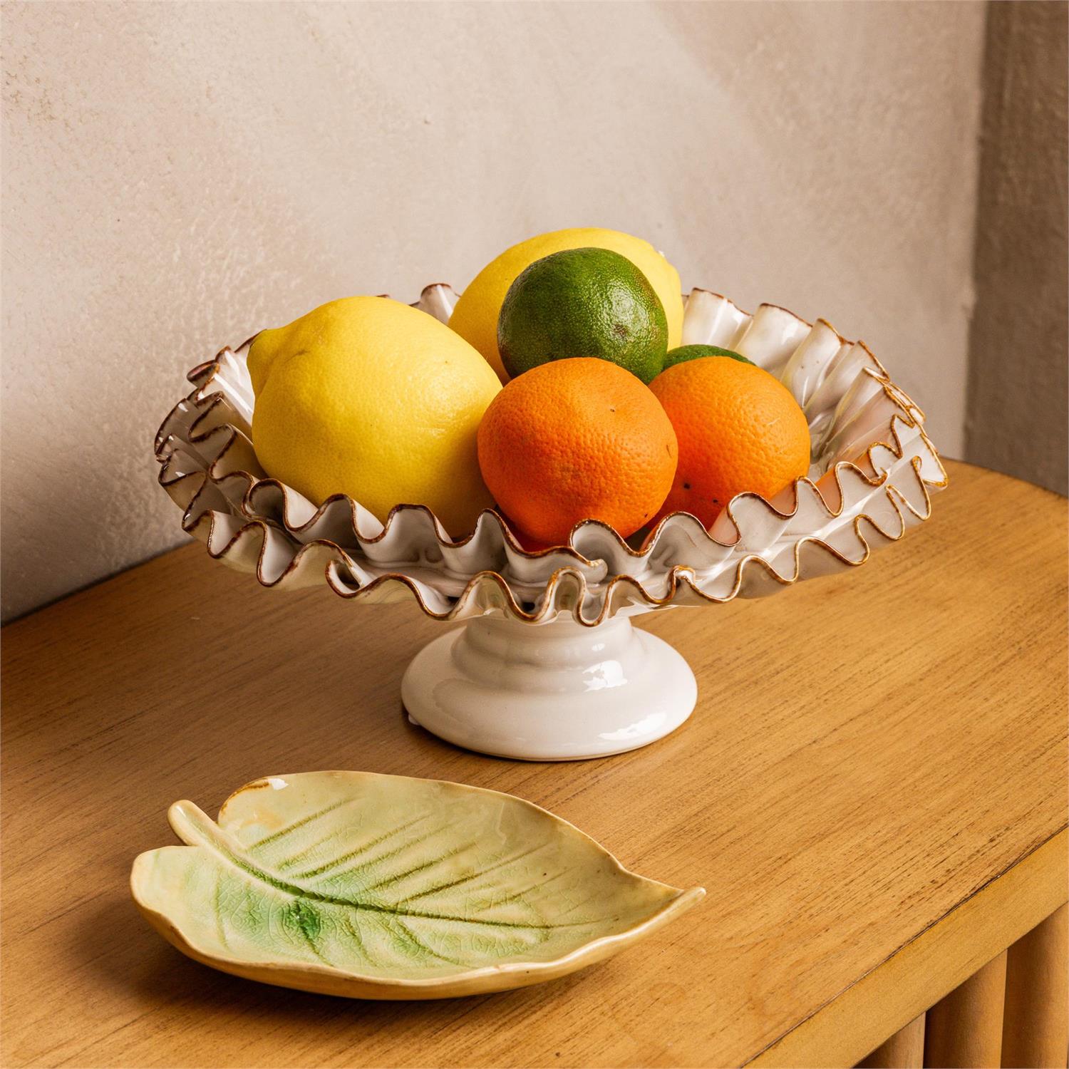 The Compote with Ruffles by Faire, featuring gold-edged white ceramic ruffles, holds lemons, limes, and oranges on a wooden table beside a ceramic leaf plate, bringing handcrafted charm to your home decor.