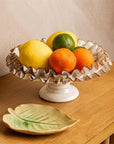 The Compote with Ruffles by Faire, featuring gold-edged white ceramic ruffles, holds lemons, limes, and oranges on a wooden table beside a ceramic leaf plate, bringing handcrafted charm to your home decor.
