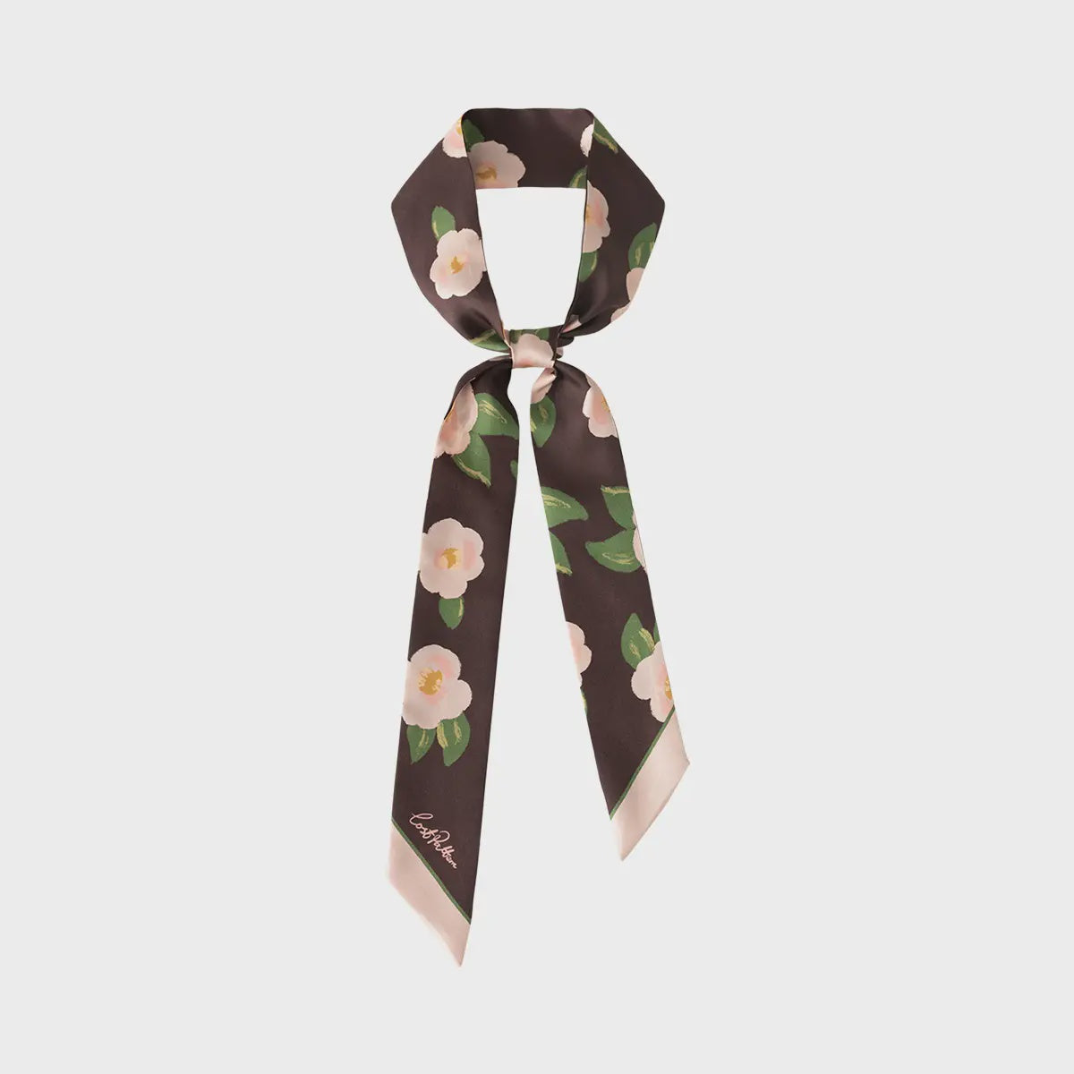 The Camellia Silk Twilly Neck Bow in Dark Brown by Faire features a floral print of white flowers and green leaves, expertly tied in a loose knot. Styled against a light gray backdrop, it exudes charm whether worn around the neck or as a chic headband.