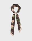 The Camellia Silk Twilly Neck Bow in Dark Brown by Faire features a floral print of white flowers and green leaves, expertly tied in a loose knot. Styled against a light gray backdrop, it exudes charm whether worn around the neck or as a chic headband.