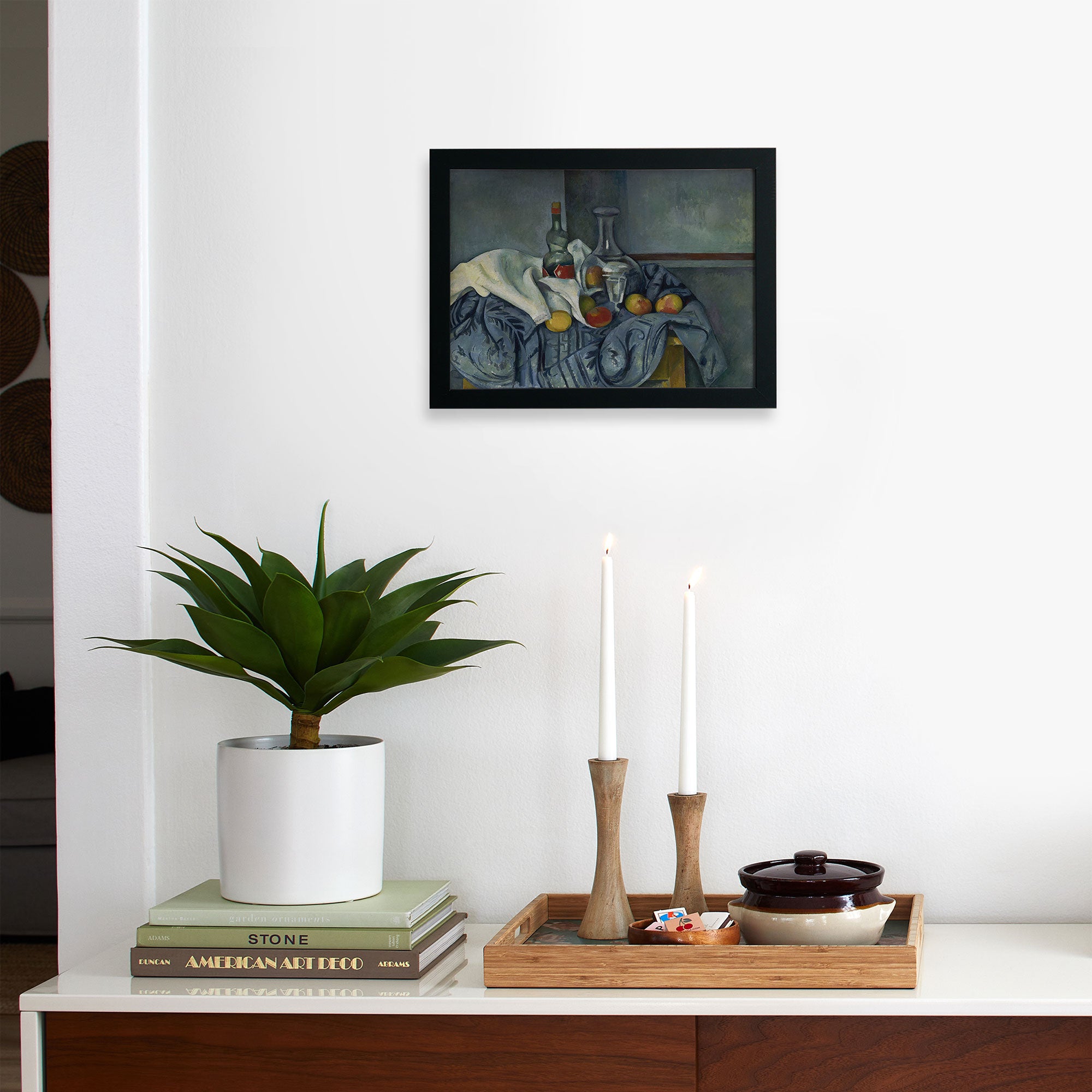 A minimalist interior displays a potted plant, stacked books, and wooden candlesticks on a tray. Above, Faire’s framed "Cezanne The Peppermint Bottle" limited-edition matte art print hangs on the white wall.