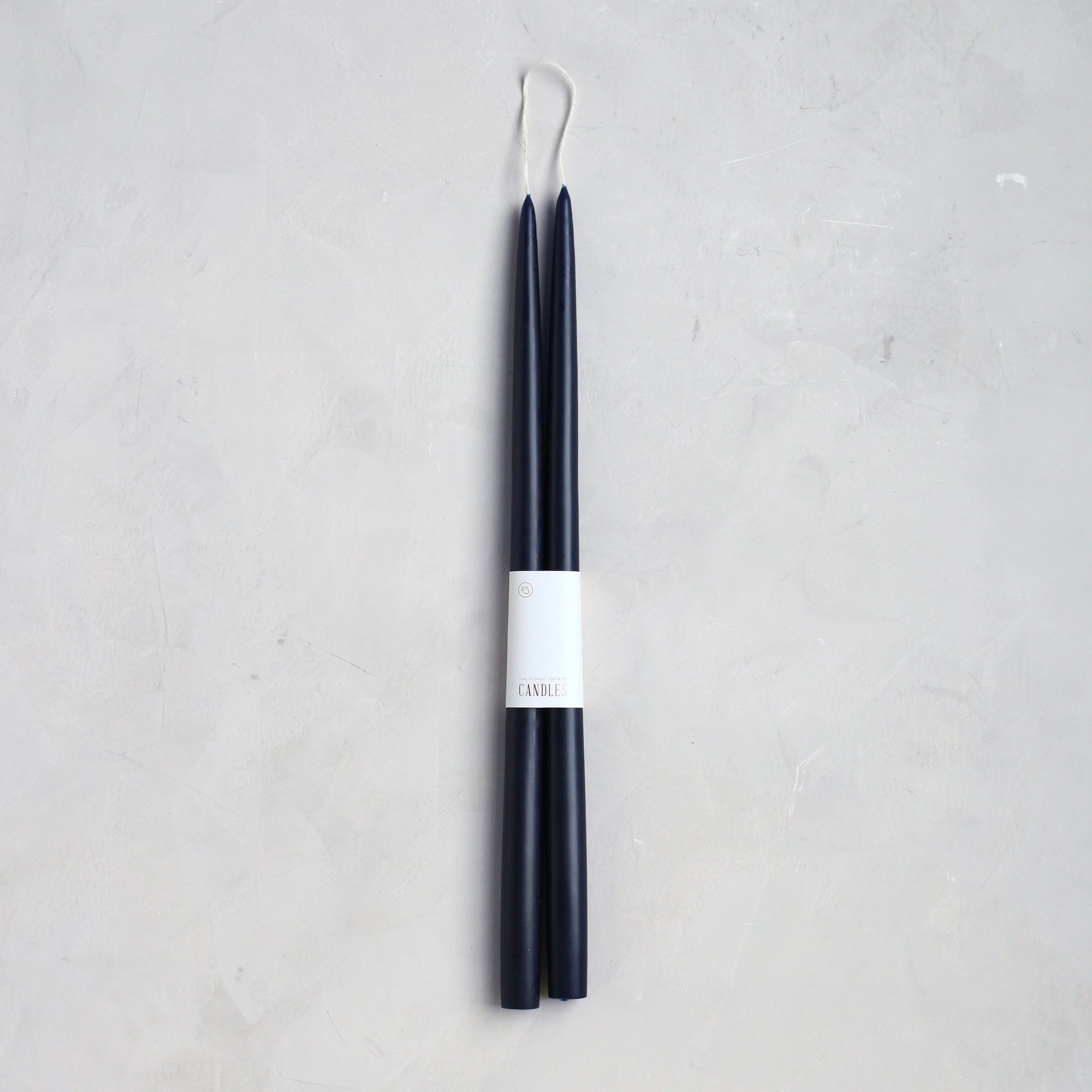 The Floral Society’s 18" Taper Candle - Pair features two tall, slender dark blue candles joined by a wick and wrapped with a white “CANDLE” label, ideal for elegant home décor. Displayed on a light gray textured surface.
