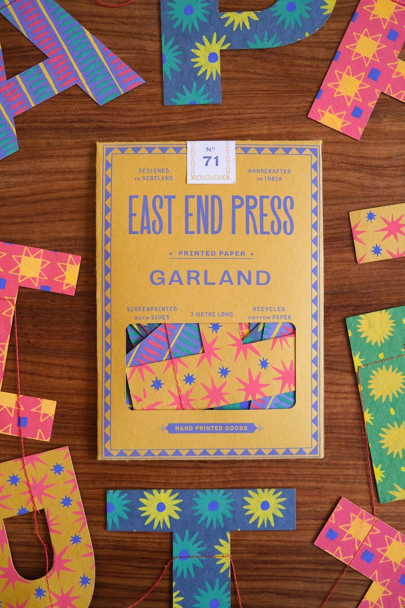 A yellow and blue Faire Paper Garland/Chains box rests on a wooden surface, surrounded by vibrant birthday garlands and paper chains in pink, blue, and yellow with floral and geometric designs.