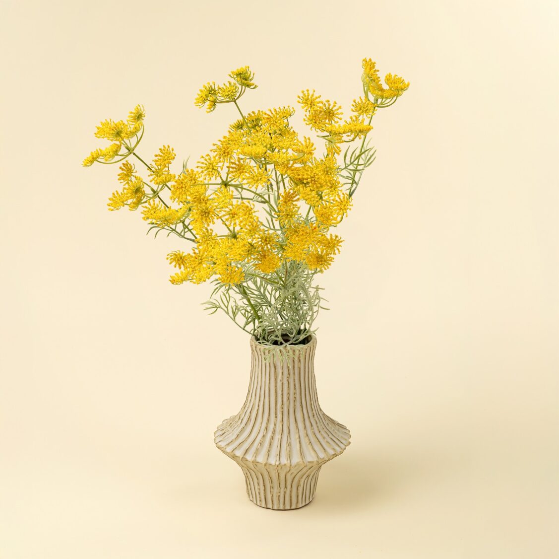 The Indaba Queen Anne Lace Spray Yellow features delicate faux flowers with thin green stems, displayed in a cream ceramic vase with vertical grooves—perfect accent decor for pale, solid backgrounds.