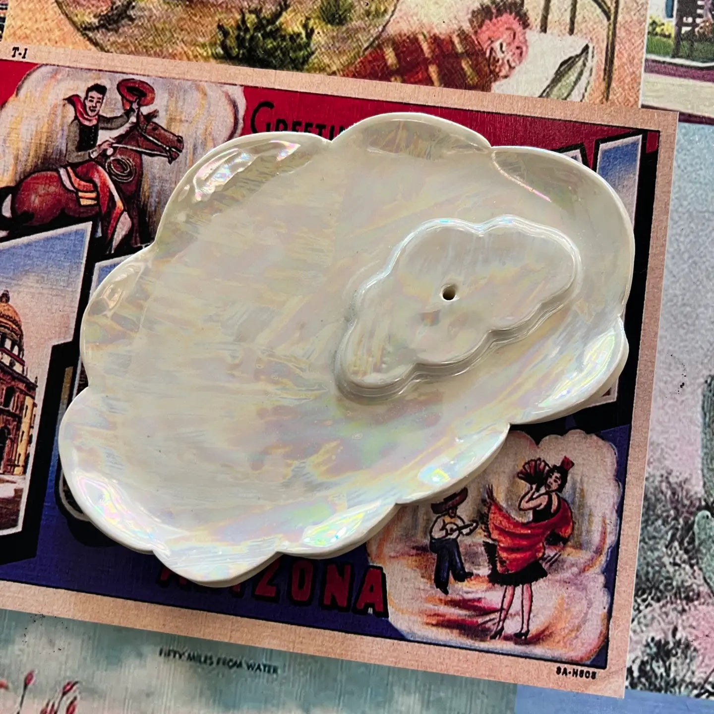 The Cloud Incense Burner by Faire features a shiny, iridescent design shaped like overlapping clouds and is displayed on a vintage postcard with colorful illustrations and text.