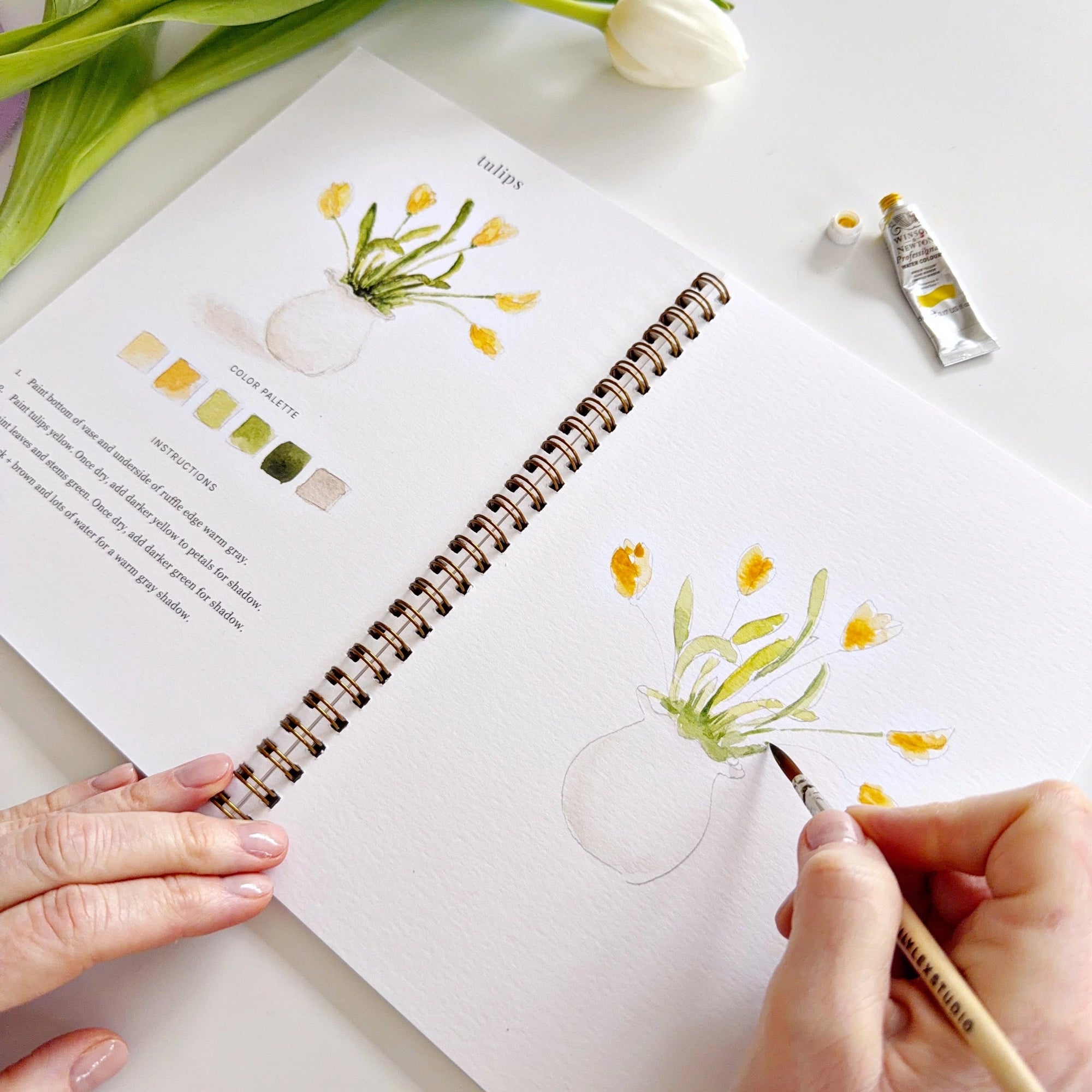 A person uses a paintbrush to paint yellow tulips in the Faire Spring Watercolor Workbook, surrounded by paint tubes, a palette, and a fresh tulip—ideal for beginner art activities on a white surface.