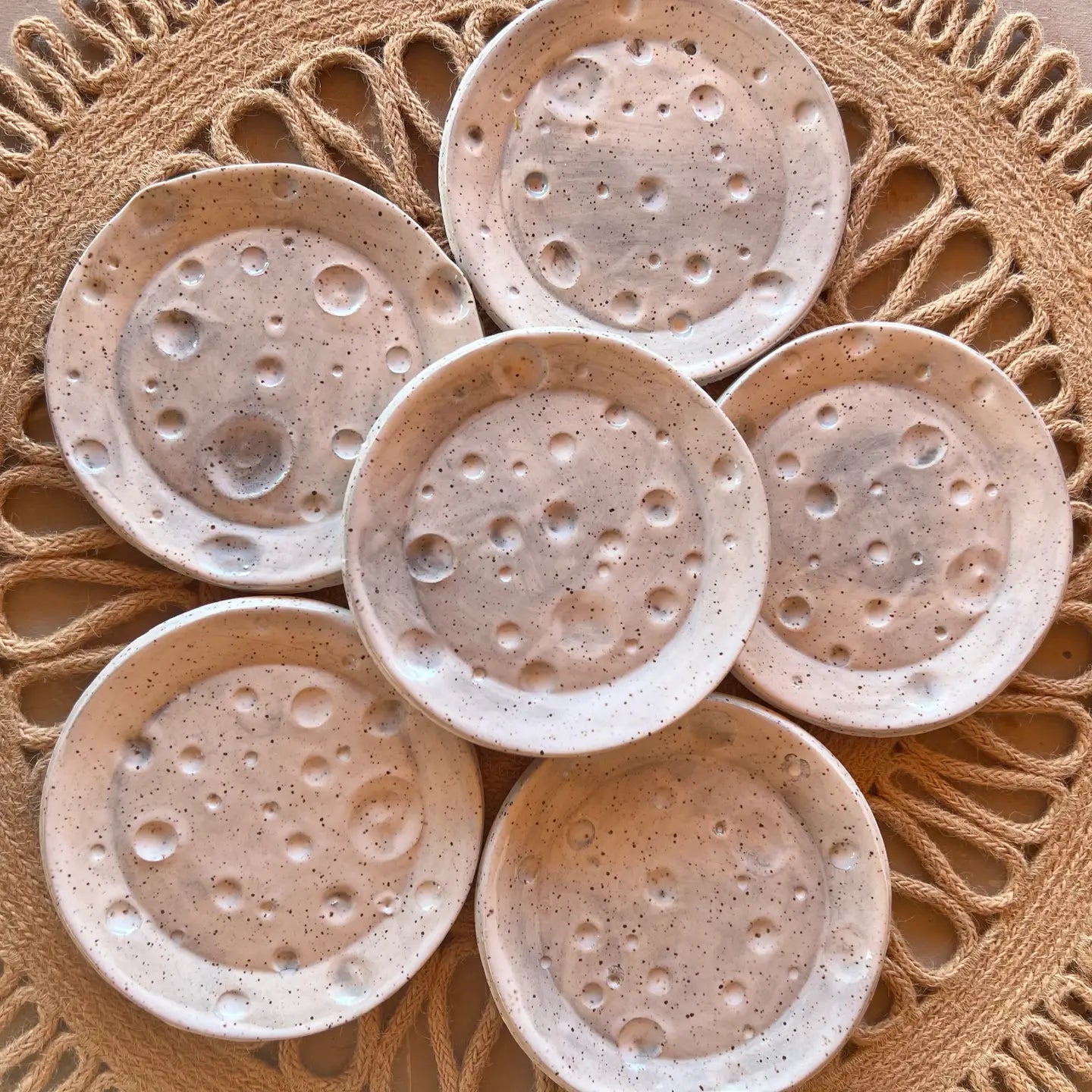 Six Moon Trinkets by Faire—small, round, speckled ceramic dishes with crater-like patterns—are arranged in a flower shape on a woven fringed circular placemat.