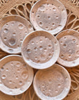 Six Moon Trinkets by Faire—small, round, speckled ceramic dishes with crater-like patterns—are arranged in a flower shape on a woven fringed circular placemat.